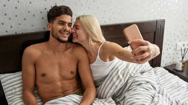 Genç, çoklu etnik çeşitlilikte gülümseyen bir çift evdeki yatakta akıllı telefondan selfie çekiyor. Beyaz kız ve Orta Doğulu adam dinleniyor ve birlikte vakit geçiriyorlar. Ev eğlencesi ve eğlence. Sabah oldu.