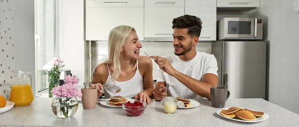 Middle eastern man feeding his caucasian girlfriend pancake during having breakfast at home. Young joyful multiracial couple spending time together. Relationship and caring. Domestic lifestyle