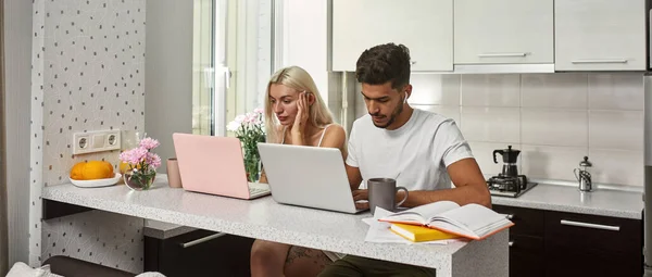 Young multiethnic couple using and working on laptops at table at home kitchen. Middle eastern man and blonde caucasian girl. Remote work and freelance. Domestic lifestyle. Modern spacious flat