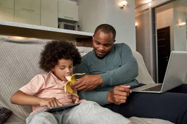 아버지는 소파에서 디지털 태블릿을 사용하는 노트북으로 아들에게 바나나를 먹이고 있습니다 — 스톡 사진