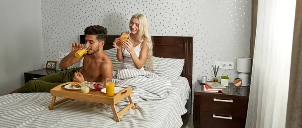 年轻的多民族夫妇在床上吃早餐 白人女孩吃三明治 中东人喝果汁 家庭生活方式 在家里休息吧现代公寓的卧室 早上时间 — 图库照片