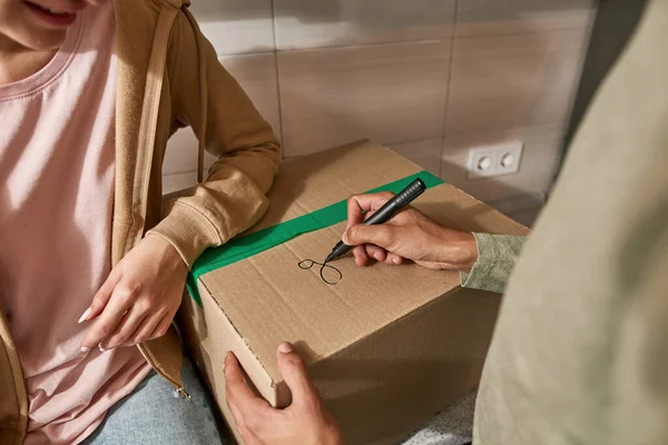 Partial of man writing g letter with marker on cardboard box near smiling girlfriend on kitchen. Young couple packing things for moving to new apartment. Concept of home relocation