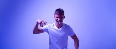 Zoomer neslinin beyaz genci kulaklıkla müzik dinlerken ve dans ederken dilini dışarı çıkartıyor. Modern genç yaşam tarzı. Mavi, beyaz ışıklı arka plan. Stüdyo çekimi. Boşluğu kopyala