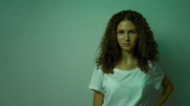 Ciddi bir beyaz kadın, elleri kalçasında kameraya bakıyor. Zoomer neslinin kıvırcık kızı tişört giyiyor. Modern genç yaşam tarzı. Yeşil arka planda izole edilmiş. Stüdyo çekimi. Boşluğu kopyala