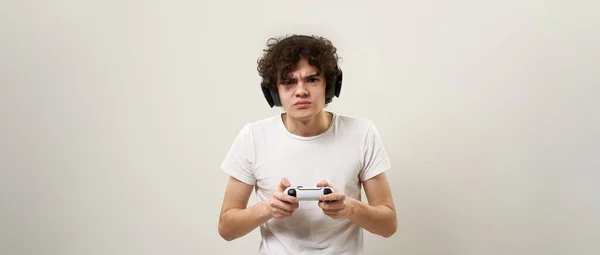 专心致志的高加索少年在操纵杆上玩电子游戏 卷曲的家伙祖传穿着T恤和耳机 现代年轻人的生活方式 白色背景 工作室拍摄 复制空间 — 图库照片