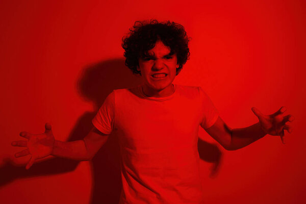 Aggressive caucasian teenage boy gesticulating with hands and looking at camera. Curly guy of zoomer generation. Concept of modern youngster lifestyle. Isolated on red background in studio. Copy space