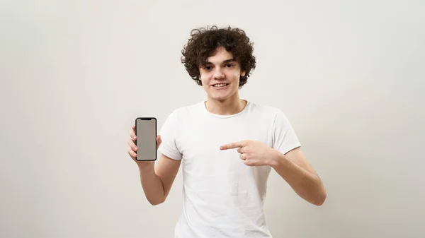 Χαμογελώντας Καυκάσιος Έφηβος Δείχνει Smartphone Και Κοιτάζοντας Την Κάμερα Σγουρομάλλης — Φωτογραφία Αρχείου