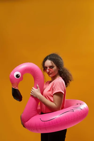 Side of european female teenage with inflatable flamingo rubber ring looking at camera. Girl of zoomer generation wearing sunglasses. Vacation and swim concept. Yellow background in studio. Copy space