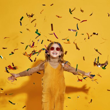 Uçan konfetilerin içinde gözlük takan beyaz, küçük yıldız kız manzarası. Zoomer kuşağının tatlı sarışın kızı. Tatil konsepti. Sarı arka planda izole edilmiş. Stüdyo çekimi