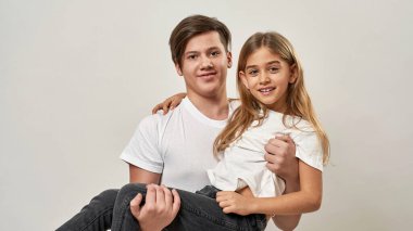 Küçük kız kardeşini kollarına alan genç bir çocuk. Gülümseyen beyaz adam ve zoomer neslinin kızı kameraya bakıyor. Modern genç yaşam tarzı. Beyaz arka planda izole edilmiş. Stüdyo çekimi. Boşluğu kopyala