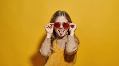 Beyaz tenli, gözlüklü, dilinden sarkan küçük bir kız şakası. Zoomer kuşağının dişi çocuğu. Çocukluk yaşam tarzı. Stüdyoda sarı arka planda izole edilmiş. Boşluğu kopyala