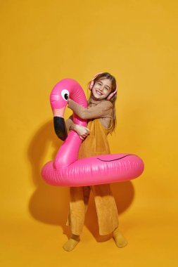 Gülümseyen küçük beyaz kız şişme flamingoya sarılıyor ve kulaklıkla müzik dinliyor. Yakınlaştırma neslinin çocuğu. Tatil ve yüzme konsepti. Stüdyoda sarı arka plan. Boşluğu kopyala