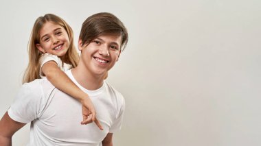 Genç çocuk kız kardeşini sırtında taşıyor. Gülümseyen beyaz adam ve zoomer neslinin kızı kameraya bakıyor. Modern genç yaşam tarzı. Beyaz arka planda izole edilmiş. Stüdyo çekimi. Boşluğu kopyala