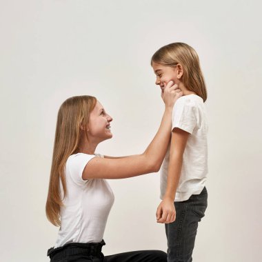 Kız kardeşinin yanaklarını çekiştiren genç bir kız. Zoomer kuşağının neşeli beyaz kız kardeşleri. Modern genç yaşam tarzı. Beyaz arka planda izole edilmiş. Stüdyo çekimi. Boşluğu kopyala