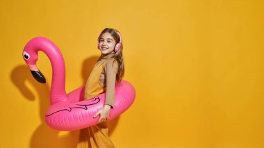 Kafkasyalı, şişme flamingo lastik halkalı, kulaklıkla müzik dinleyen bir kızın yan görüntüsü. Zoomer kuşağının çocuğu. Tatil ve yüzme konsepti. Sarı arka plan. Stüdyo çekimi. Boşluğu kopyala