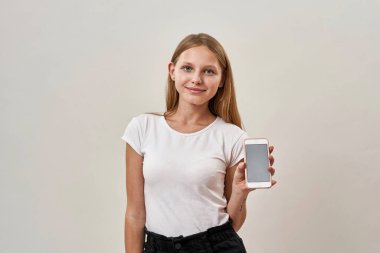 Gülümseyen beyaz kız cep telefonunu gösteriyor ve kameraya bakıyor. Zoomer kuşağının sarışın kızı. Modern genç yaşam tarzı. Beyaz arka planda izole edilmiş. Stüdyo çekimi. Boşluğu kopyala