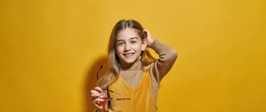 Gözlüklü, gülümseyen beyaz bir kız ve kameraya bakıyor. Zoomer kuşağının dişi çocuğunun kırpılmış görüntüsü. Modern çocukluk tarzı. Stüdyoda sarı arka planda izole edilmiş. Boşluğu kopyala