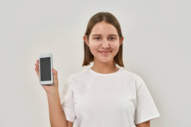 Gülümseyen beyaz kız akıllı telefon gösteriyor ve kameraya bakıyor. Zoomer kuşağının kadın tişörtü. Modern gençlik yaşam tarzı kavramı. Beyaz arka planda izole edilmiş. Stüdyo çekimi