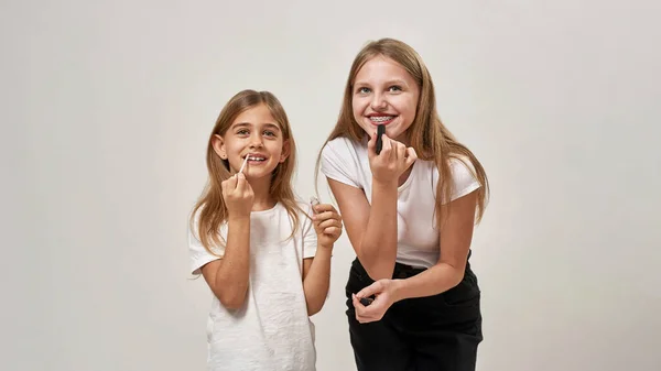 Teenage and little girls painting lips with lipsticks. European sisters of zoomer generation looking away. Modern youngster lifestyle. Female beauty. Isolated on white background in studio. Copy space