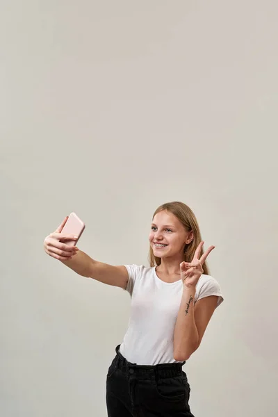 Uśmiechnięta Biała Nastolatka Pokazuje Znak Zwycięstwa Podczas Robienia Selfie Smartfonie — Zdjęcie stockowe