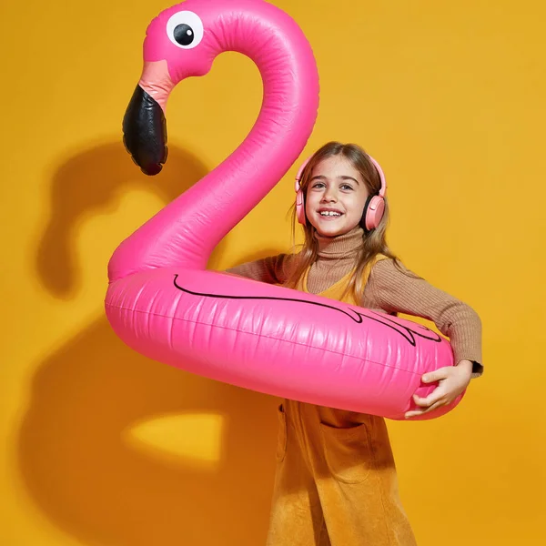 Smiling caucasian little girl with inflatable flamingo rubber ring listening music in headphones. Female child of zoomer generation. Vacation and swim concept. Yellow background in studio. Copy space