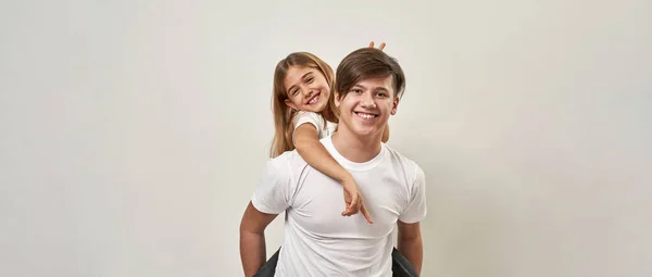Teenage boy holding little sister piggyback while she making horns sign over his head. Cheerful european guy and girl of zoomer generation looking at camera. White background. Studio shoot. Copy space