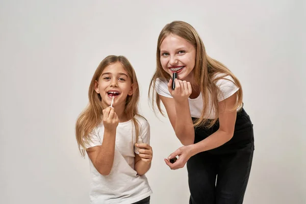 Teenage and little girls painting lips with lipsticks. Smiling caucasian sisters of zoomer generation look at camera. Modern youngster lifestyle. Female beauty. White background in studio. Copy space