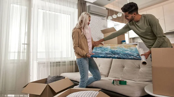 Side view of young multiethnic couple packing picture with stretch film at studio flat. Blonde caucasian girl and smiling bearded middle eastern man moving to new apartment. Concept of home relocation