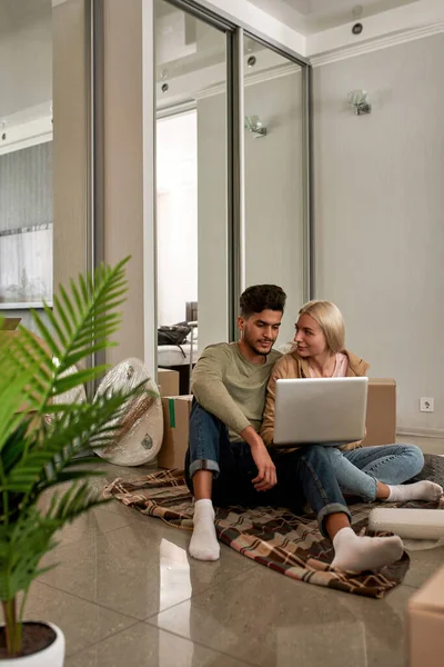 Young multiethnic couple using and watching laptop on blanket on floor with cardboard boxes and things. Caucasian girl and middle eastern man move to new apartment. Concept of home relocation