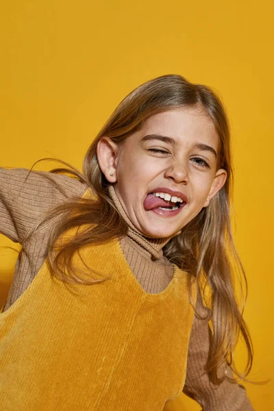 Cropped image of joking caucasian little girl winking and sticking out tongue. Cute female kid of zoomer generation. Concept of modern childhood lifestyle. Isolated on yellow background. Studio shoot