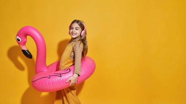 Side view of smiling caucasian girl with inflatable flamingo rubber ring listening music in headphones. Kid of zoomer generation. Vacation and swim concept. Yellow background. Studio shoot. Copy space
