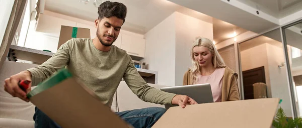 Bottom view of middle eastern man looking at open cardboard box near caucasian girl using laptop on sofa in spacious flat. Young multiethnic couple moving to their new apartment. Home relocation