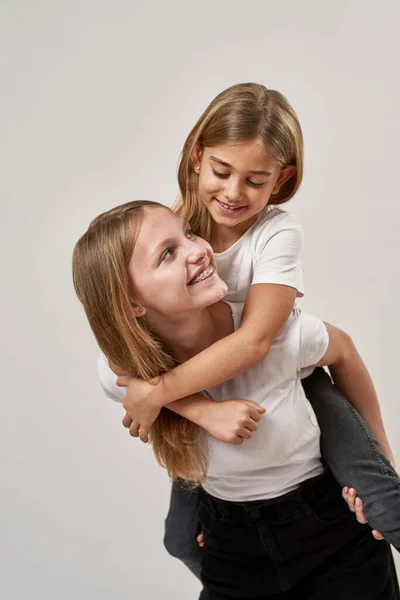Teenage girl holding little sister piggyback. Smiling caucasian sisters of zoomer generation looking at each other. Modern youngster lifestyle. Isolated on white background. Studio shoot. Copy space
