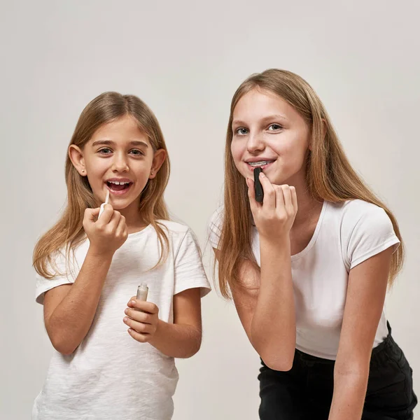 Teenage and little girls painting lips with lipsticks. Blonde smiling european sisters of zoomer generation looking at camera. Modern youngster lifestyle. Female beauty. White background. Studio shoot