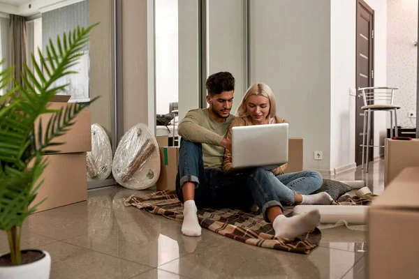 Young concentrated multiracial couple using laptop on blanket on floor with cardboard boxes and things for packing. Caucasian girl and middle eastern man move to new apartment. Home relocation