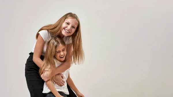 Teenage girl hugging her little sister and looking at camera. Cheerful caucasian sisters of zoomer generation enjoying time together. Modern youngster lifestyle. White background in studio. Copy space