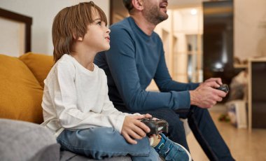 Kafkasyalı küçük bir çocuk ve sakat bir baba evdeki kanepede joysticklerle oyun oynuyorlar. Odaklanmış çocuk ve erkek birlikte vakit geçiriyorlar. Babalık ve ilişki. Ev içi eğlence ve eğlence.