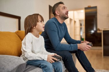 Avrupalı küçük çocuk ve babası evdeki kanepede joysticklerle oyun oynuyorlar. Konsantre çocuk ve ağzı açık adam birlikte zaman geçiriyorlar. Babalık ve ebeveynlik. Boş zaman ve eğlence.