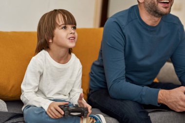 Kafkasyalı küçük çocuk ve kısmi baba evdeki kanepede joysticklerle oyun oynuyorlar. Konsantre çocuk ve erkek birlikte zaman geçiriyorlar. Babalık ve ebeveynlik. Ev içi eğlence ve eğlence.