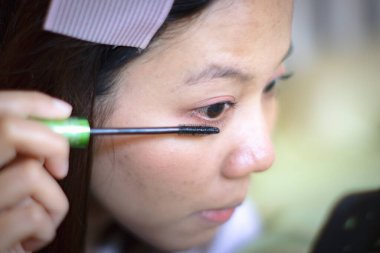 Güzel Asain kadını aynanın önünde makyaj yapıyor (kirpiklerini fırçalıyor ve boyuyor). Seçici odak.