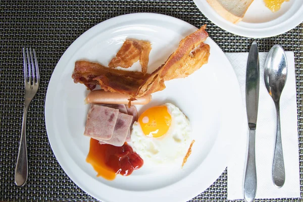 American breakfast with fried eggs, bacon, ham, and sausage on a white plate.