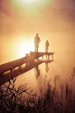 Göl kenarında güzel bir sabah sahnesi. Baba ve oğul güneşin doğuşunda balık tutuyor..