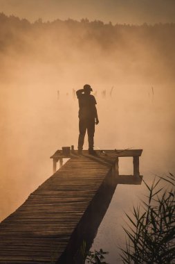 Göl kenarında güzel bir sabah sahnesi. Güneşin doğuşunda tahta bir iskelede balık tutan bir adamın silueti..