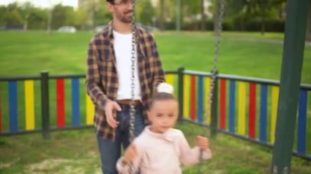 Мужчина играет со своей приемной дочерью на качелях на детской площадке. — стоковое видео