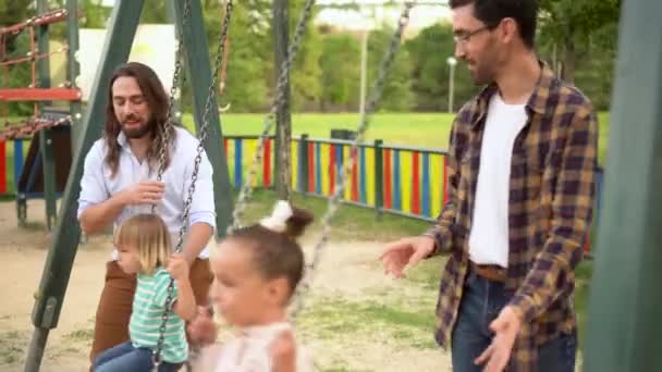 Ein junges schwules Paar spielt mit seinen Kindern auf der Schaukel auf dem Spielplatz. — Stockvideo