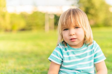 Çocuk parktaki çimlere oturmuş kameraya bakıyor.