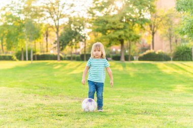 Küçük bir çocuk kameraya bakıyor, topla oynuyor.