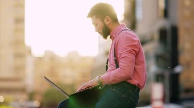 Arka planda şehir manzarası olan Asyalı bir adam dışarıda dizüstü bilgisayarla çalışıyor.