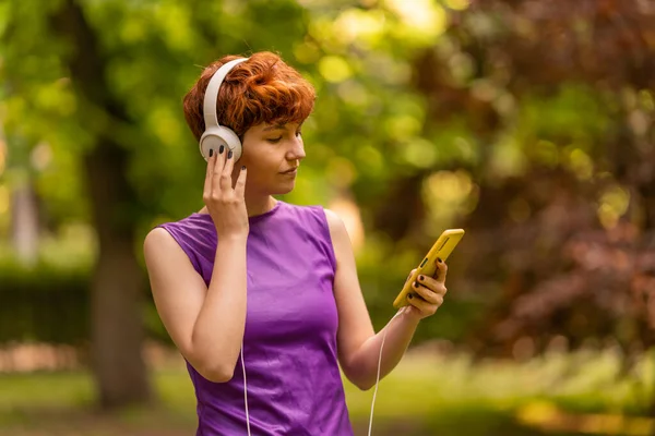 Non binary person in purple top with red hair touching headphones and browsing playlist on cellphone while listening to music on sunny day in park