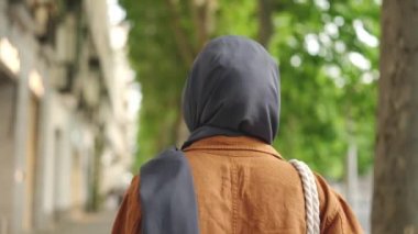 Başörtüsü içinde Müslüman bir kadın sokakta yürüyor kameraya gülümsüyor ve yürümeye devam ediyor.
